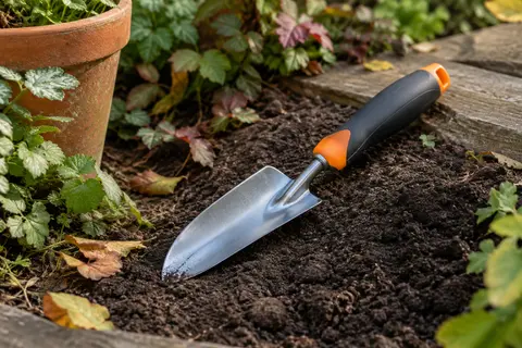 Déplantoir à tête en acier et poignée noire et orange posé près d’une terre de jardin ameublie.