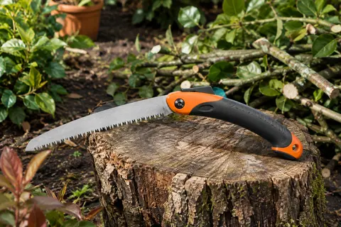 Scie pliante d’élagage noire et orange à lame dentée posée sur une souche dans un jardin