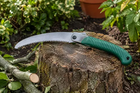 Scie pliante d’élagage à poignée verte et lame incurvée posée sur du bois dans un jardin