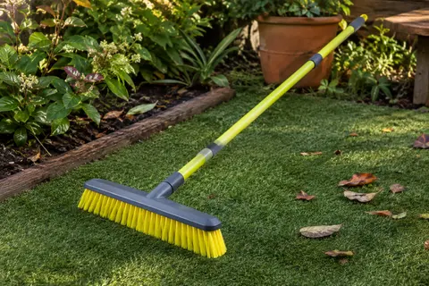 Brosse à poils coudés et manche extensible posée sur du gazon artificiel avec quelques feuilles