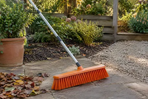 Balai-brosse TreeLen orange à tête large posé sur un patio de jardin près de feuilles balayées.
